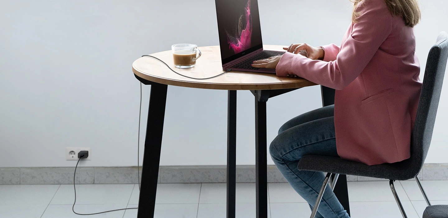 Человек сидит за столом и пользуется ноутбуком Galaxy Book. USB-кабель подключён к ноутбуку, а другой его конец — к розетке через адаптер. Кабель провисает благодаря своей большой длине.