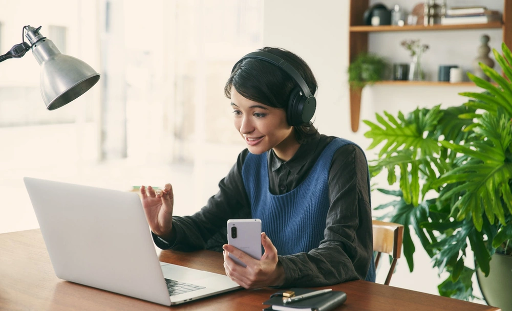 Женщина за столом в наушниках Sony WH-CH720N пользуется ноутбуком и смартфоном